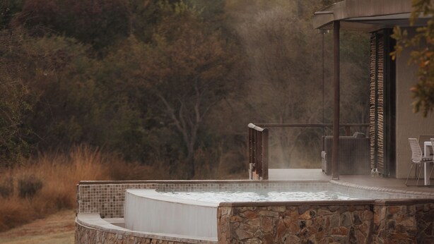 A stone-built hot tub with a tiled edge, surrounded by a stone wall, set against a backdrop of trees and hills. - Babirwa Game Ranch (Rooiberg)
