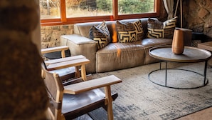 A cozy living room with a leather sofa, wooden chairs, a round coffee table, and a stone wall. - Babirwa Game Ranch (Rooiberg)
