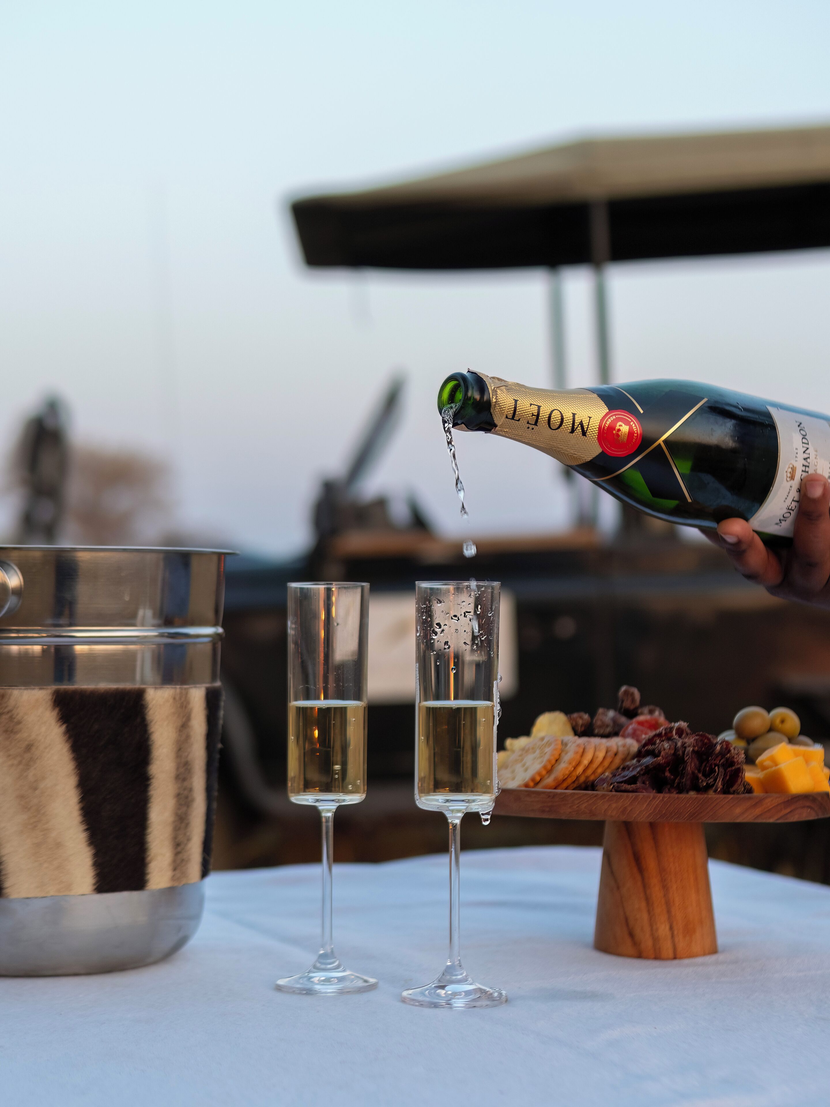 A bottle of Moët & Chandon champagne being poured into two glasses, with a wooden board of cheeses and crackers in the background.