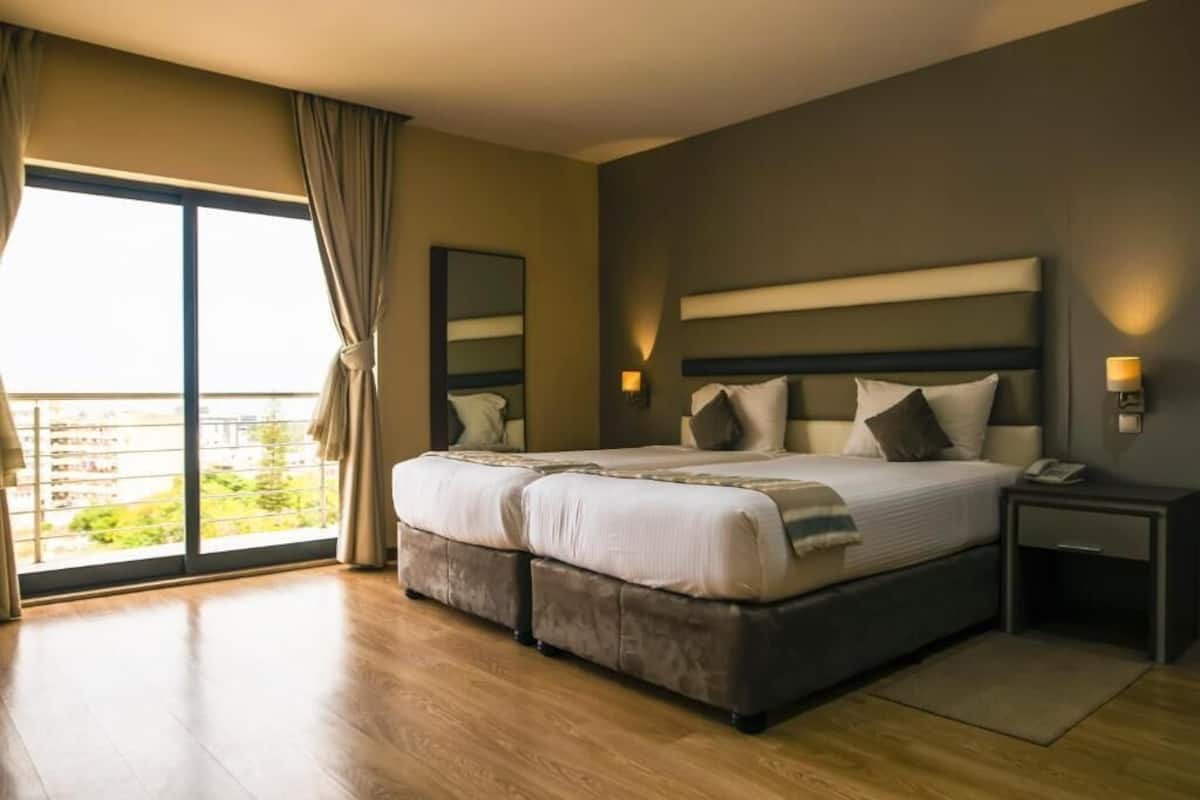 A double bed with a headboard, bedside tables, and a view of greenery through large windows.