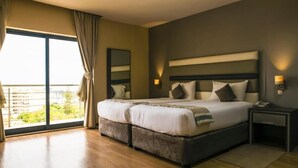 A double bed with a headboard, bedside tables, and a view of greenery through large windows. - WL Hotel Maputo City Center (Maputo)