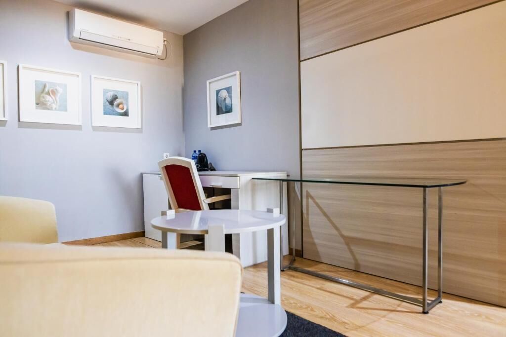 A modern hotel room with a bed, a desk, a chair, and a glass table. There are framed pictures on the wall and an air conditioning unit above the desk.