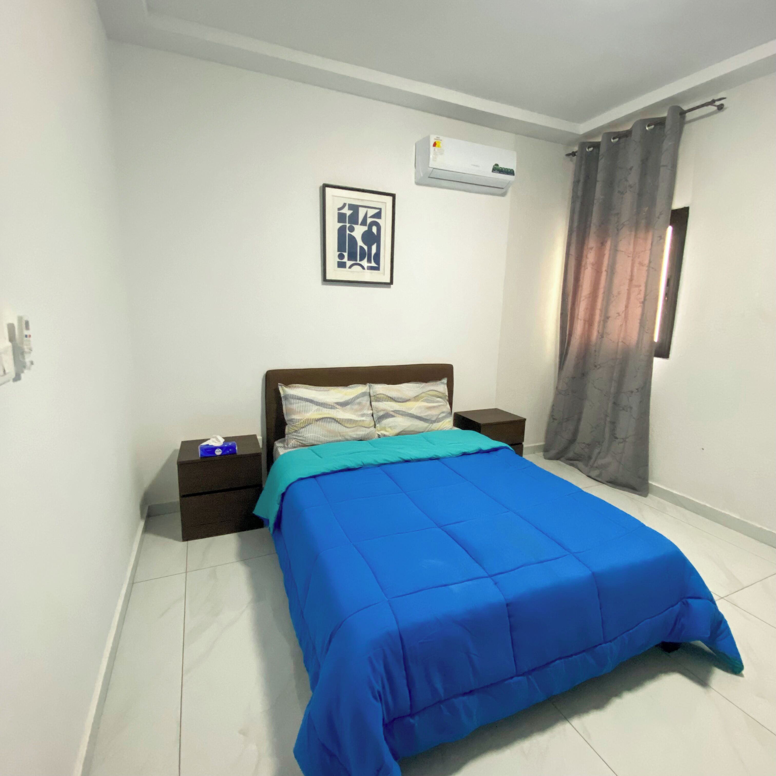 A bedroom with a bed, a nightstand, a wall-mounted air conditioner, a framed picture, and a window with curtains.