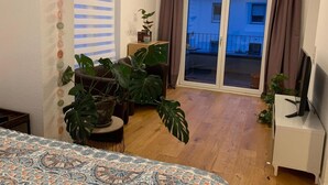 Huoneessa on sänky, televisio, parveke, jolta on näkymä, ja suuria huonekasveja.