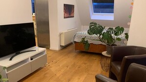Moderni makuuhuone, jossa on sänky, jalustalla oleva televisio, ruukkukasvi ja ikkuna.