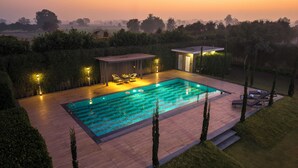 A modern outdoor swimming pool area with a wooden deck, lounge chairs, and a covered seating area. - Elivaas Levante Farms 5 BHK Villa (New Delhi)