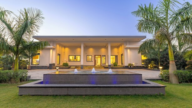 A modern building with a large entrance, a water feature in front, and palm trees around. - Elivaas Levante Farms 5 BHK Villa (New Delhi)