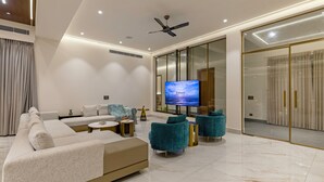 A modern living room with a large flat-screen TV, a sofa, teal armchairs, and a coffee table. - Elivaas Levante Farms 5 BHK Villa (New Delhi)