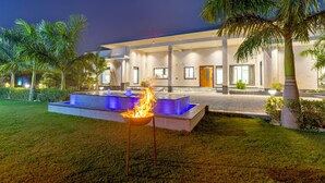 A modern house with a pool, a fire feature, and palm trees. - Elivaas Levante Farms 5 BHK Villa (New Delhi)