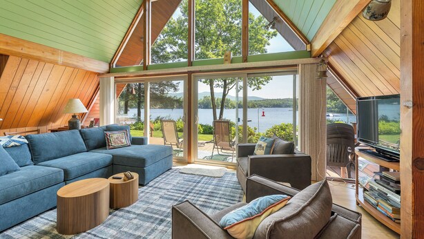 Una sala de estar acogedora con un amplio ventanal con vistas a un lago, un sofá seccional, una mesa de centro y una televisión.