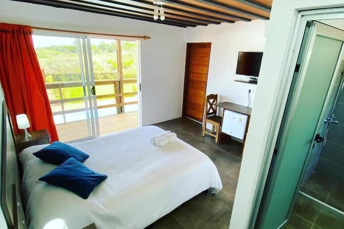A bedroom with a bed, a desk, a TV, and a door leading to a balcony.