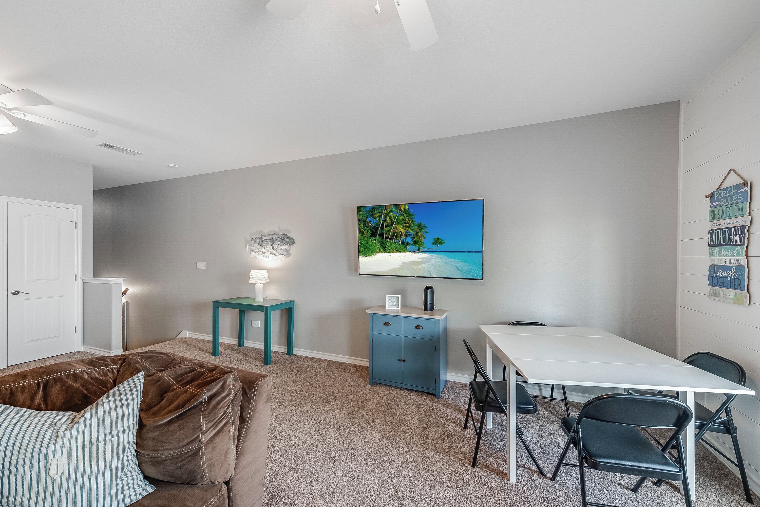Een kamer met een witte plafondventilator, een blauwgroene bijzettafel met een lamp, een blauwe kast, een witte tafel met vier stoelen en een schilderij van een strand.