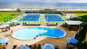 A pool area with a tennis court, lounge chairs, and umbrellas.