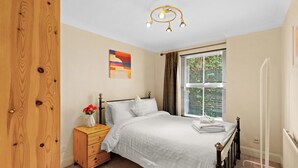 A bedroom with a wooden wardrobe, a bedside table with a vase of flowers, a bed with white bedding, a window with curtains, and a ceiling light fixture. - 2bed 2bath close to Tower Bridge (London)