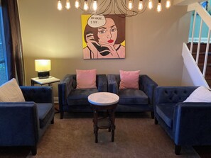 Ein Wohnzimmer mit zwei dunkelblauen Sofas, einem runden Couchtisch und einer Lampe auf einem Beistelltisch. Ein großes Gemälde mit Sprechblase ist an der Wand.