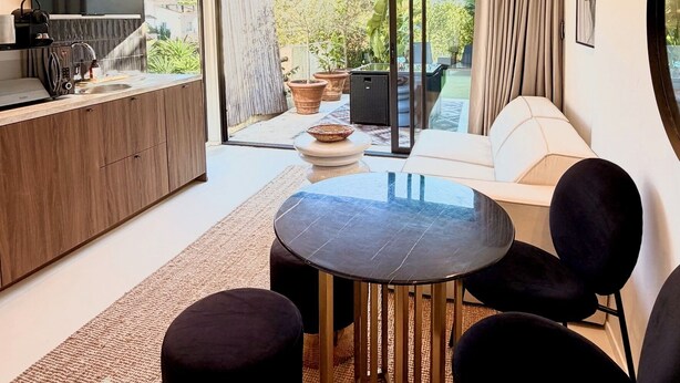 Una sala de estar moderna con una mesa redonda, sillas negras, un sofá y una zona de cocina con televisor y mostrador.
