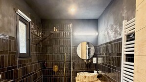 Un baño pequeño con paredes de azulejos oscuros, un tocador de madera, un espejo redondo y una cabina de ducha.