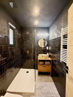 Ein kleines Badezimmer mit dunkel gefliesten Wänden, einem Holzkosmetikschrank, einem runden Spiegel und einer Duschkabine.
