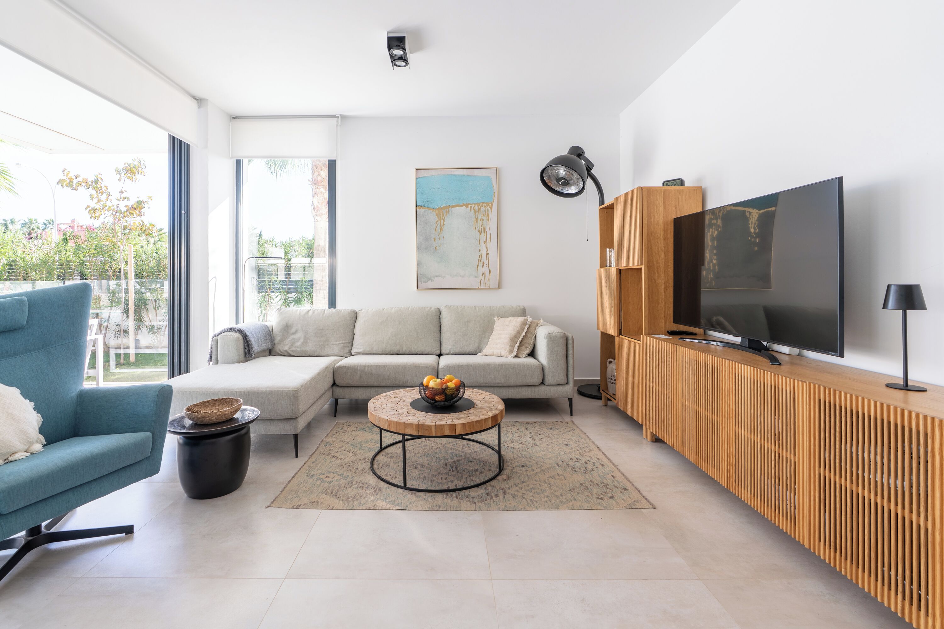 Una sala de estar moderna con un sofá, un sillón azul, una mesa redonda de centro con fruta, un televisor y un mueble bajo de madera.