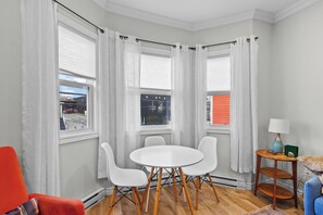A corner room with a round table and four chairs, large windows with white curtains, a small table with a lamp, and a red chair. - Cozy 2BR Central Home | 5 Min to Downtown & George (St. John's)