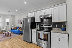 A modern kitchen with white cabinets, stainless steel appliances, and a blue sofa in the adjacent living area. - Cozy 2BR Central Home | 5 Min to Downtown & George (St. John's)