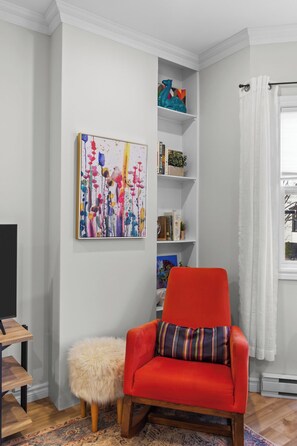 A modern living room with a red armchair, a fluffy ottoman, a bookshelf, and a colorful abstract painting. - Cozy 2BR Central Home | 5 Min to Downtown & George (St. John's)