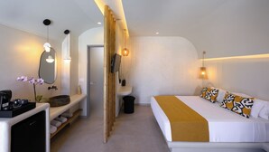 A modern hotel room with a bed, bedside table, mirror, and a small seating area with a bench and a potted plant. - Anemone Luxury Suites (Santorini)