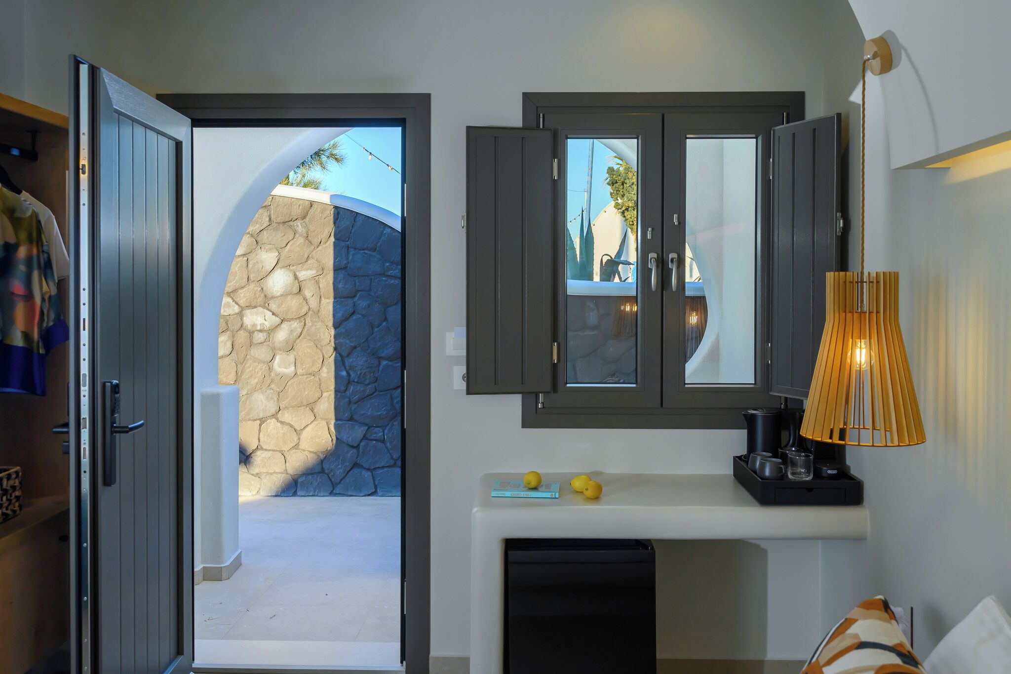 A hotel room with a stone wall, a wardrobe, a mirror, a lamp, and a small table with a book and lemons.