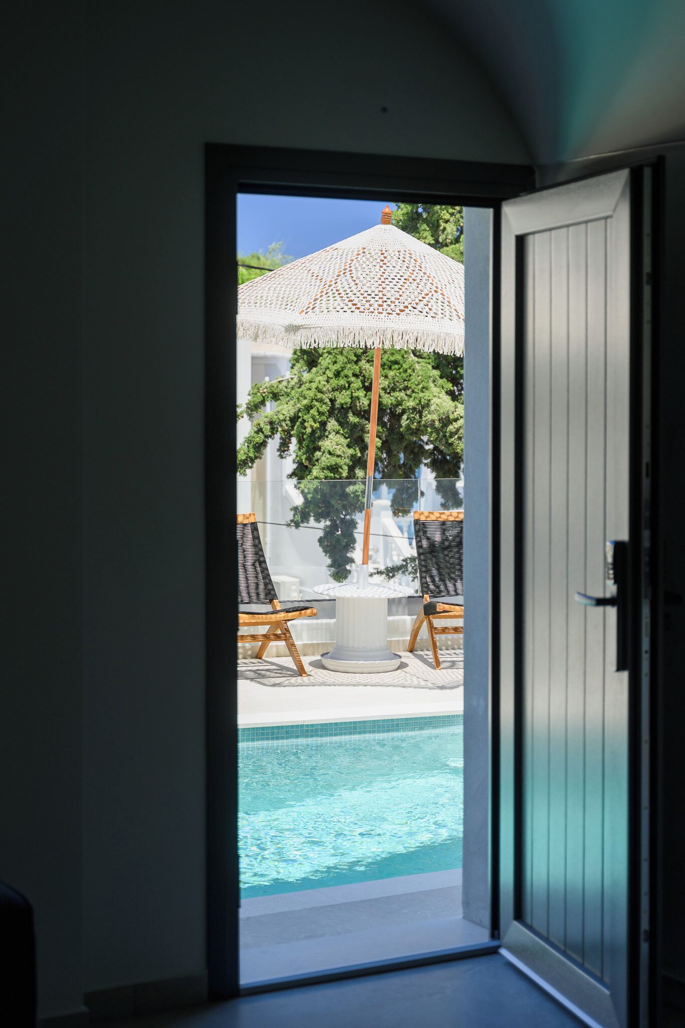 An open door leading to a pool area with a white umbrella, two lounge chairs, and a small table.