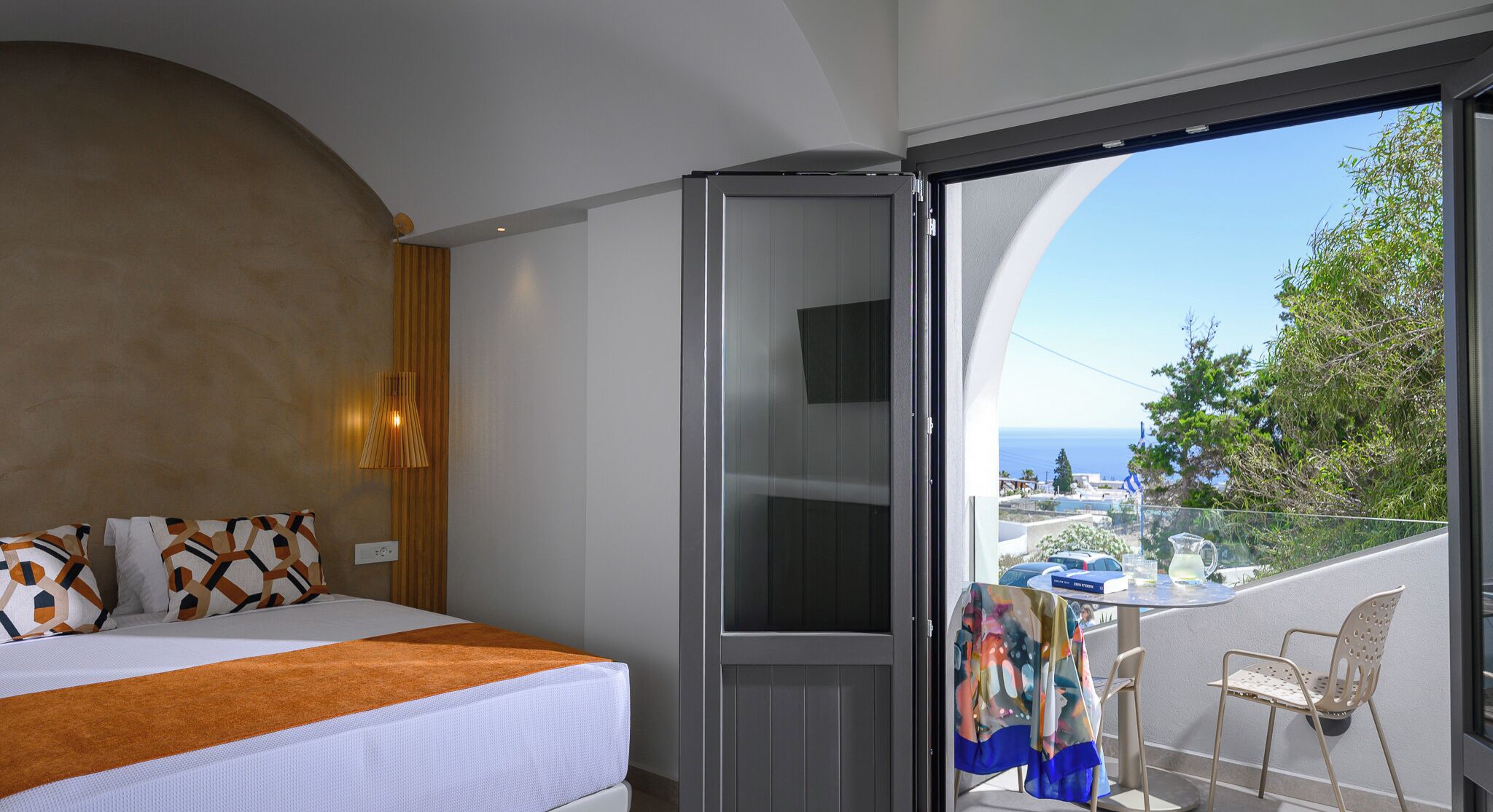 A bedroom with a bed, a TV, a balcony with a table and chairs, and a view of the sea.