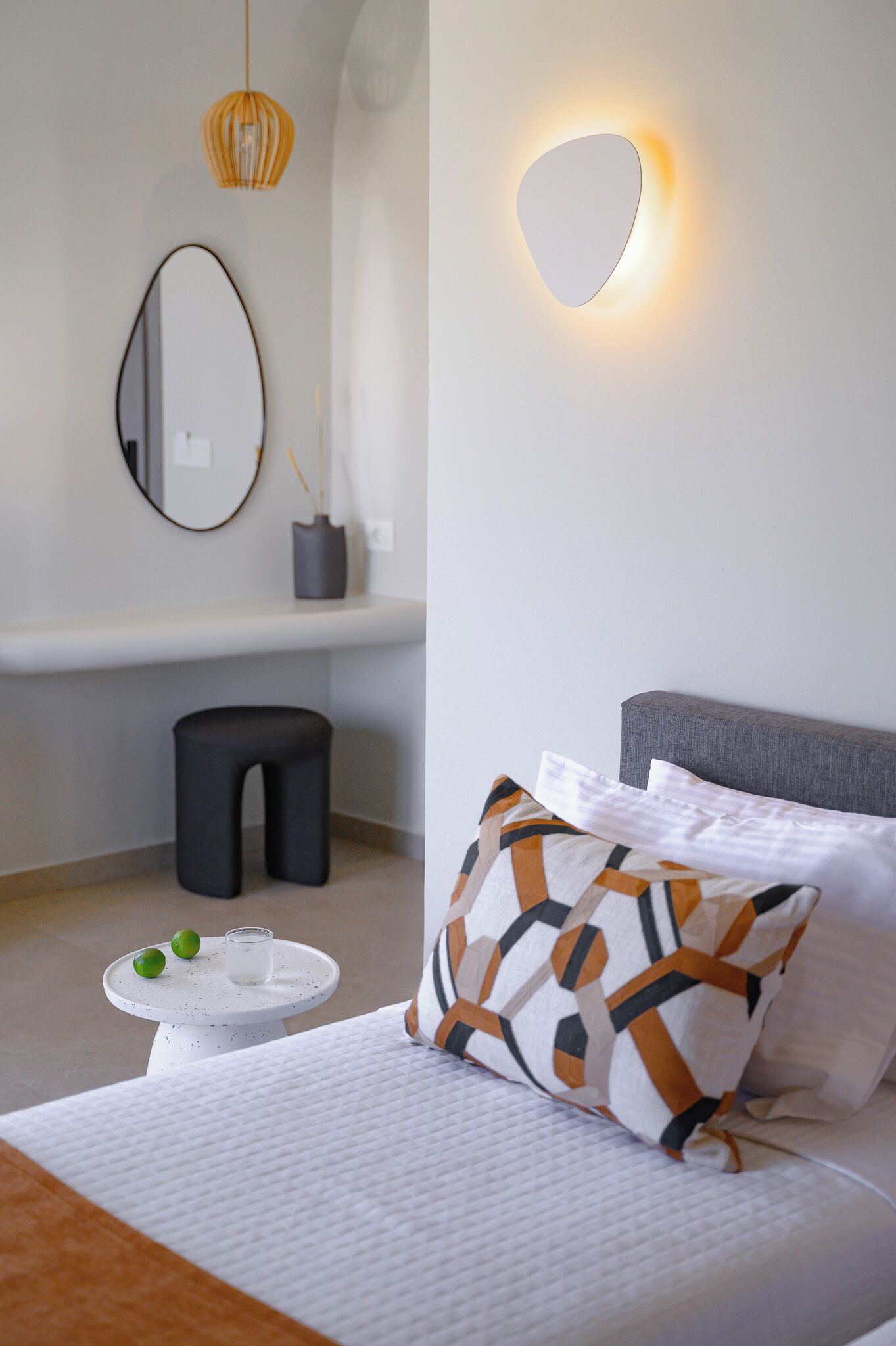 A modern bedroom with a bed, a bedside table with a glass and limes, a round mirror, and a wall-mounted light.