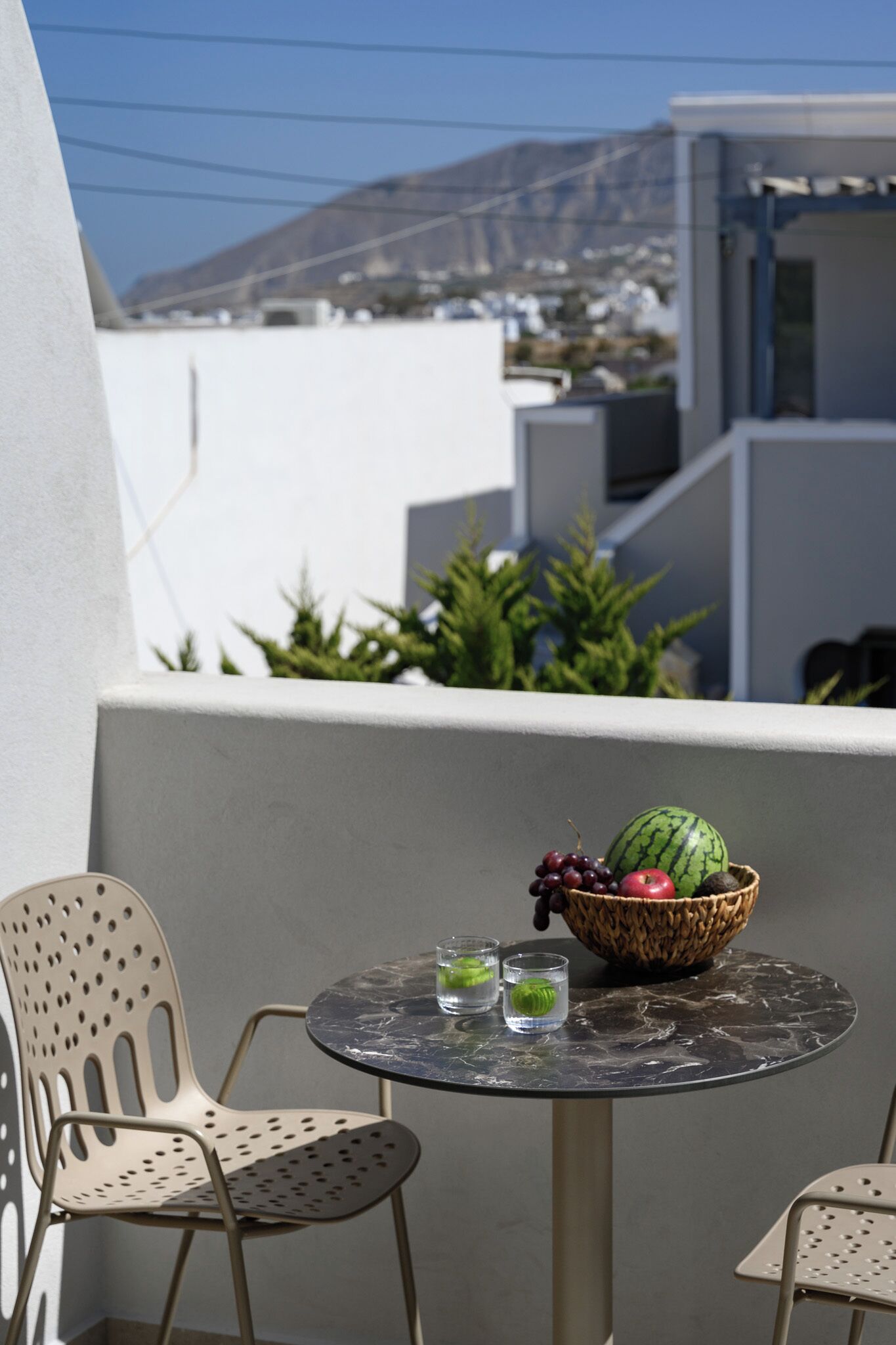 A balcony with a table, chairs, a bowl of fruit, and a view of buildings and a mountain.