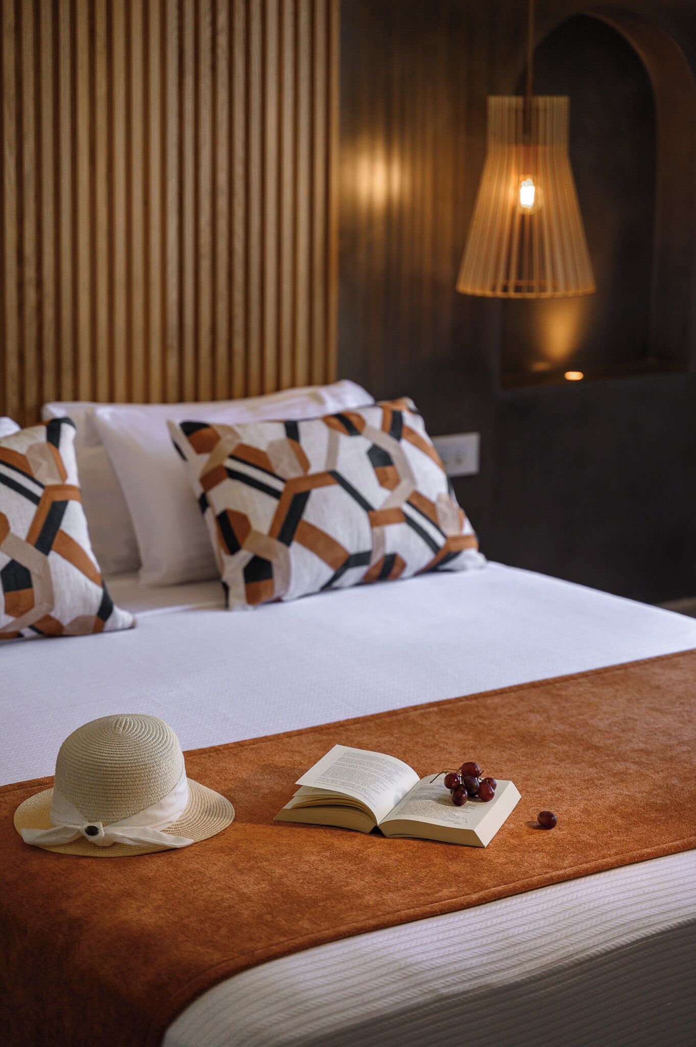 A bed with a white comforter, a brown throw blanket, a straw hat, an open book, and a bunch of grapes on the bed.
