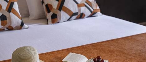 A bed with a white comforter, a brown throw blanket, a straw hat, an open book, and a bunch of grapes on the bed.