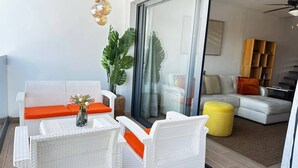 Ein modernes Wohnzimmer mit weißen Rattanmöbeln, einem gelben Ottomanen und einer Glastür, die auf einen Balkon führt.
