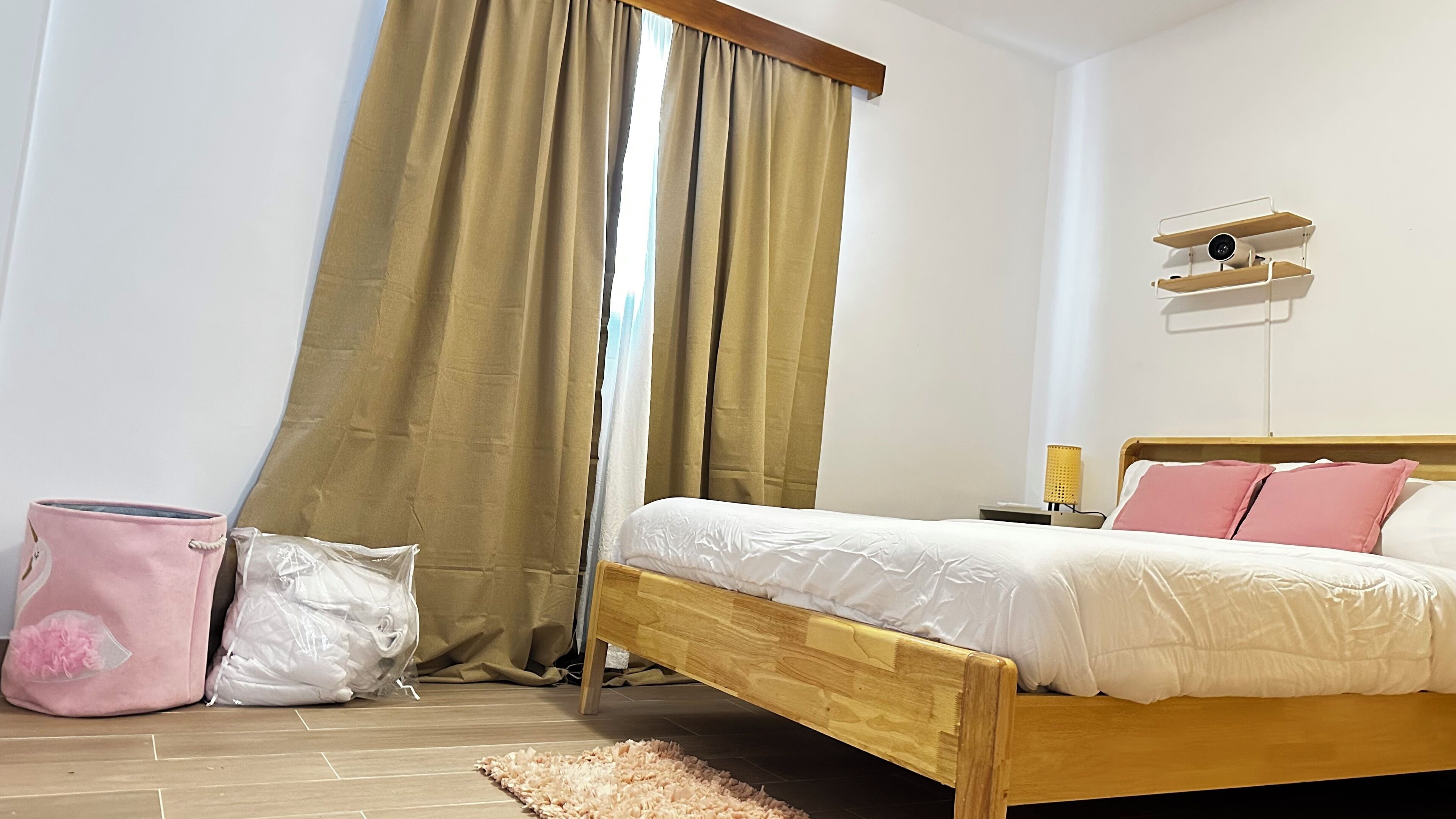 Una camera da letto con un letto di legno, tende beige, un comodino con una lampada e una mensola a parete con una fotocamera.