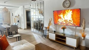 Ein modernes Wohnzimmer mit einem großen Flachbildfernseher, einer Couch, einem Couchtisch und einem Essbereich im Hintergrund.
