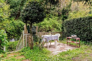 Um pátio com jardim, mesa e cadeiras, cercado por vegetação exuberante e um muro de madeira.