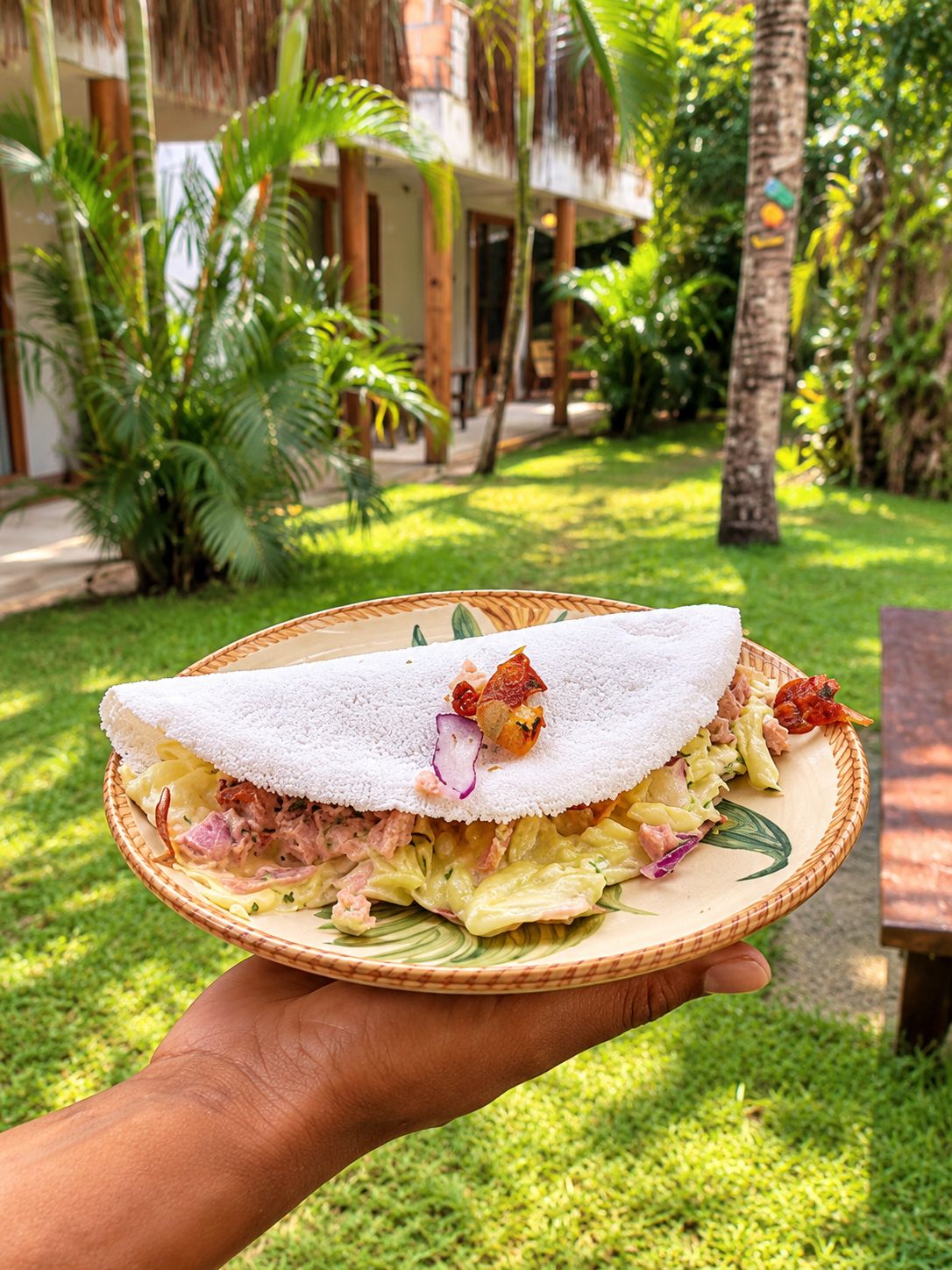 A person holding a plate with a rolled food item, possibly a crepe, garnished with a piece of meat and a red vegetable, set against a lush green garden with palm trees and a building in the background.