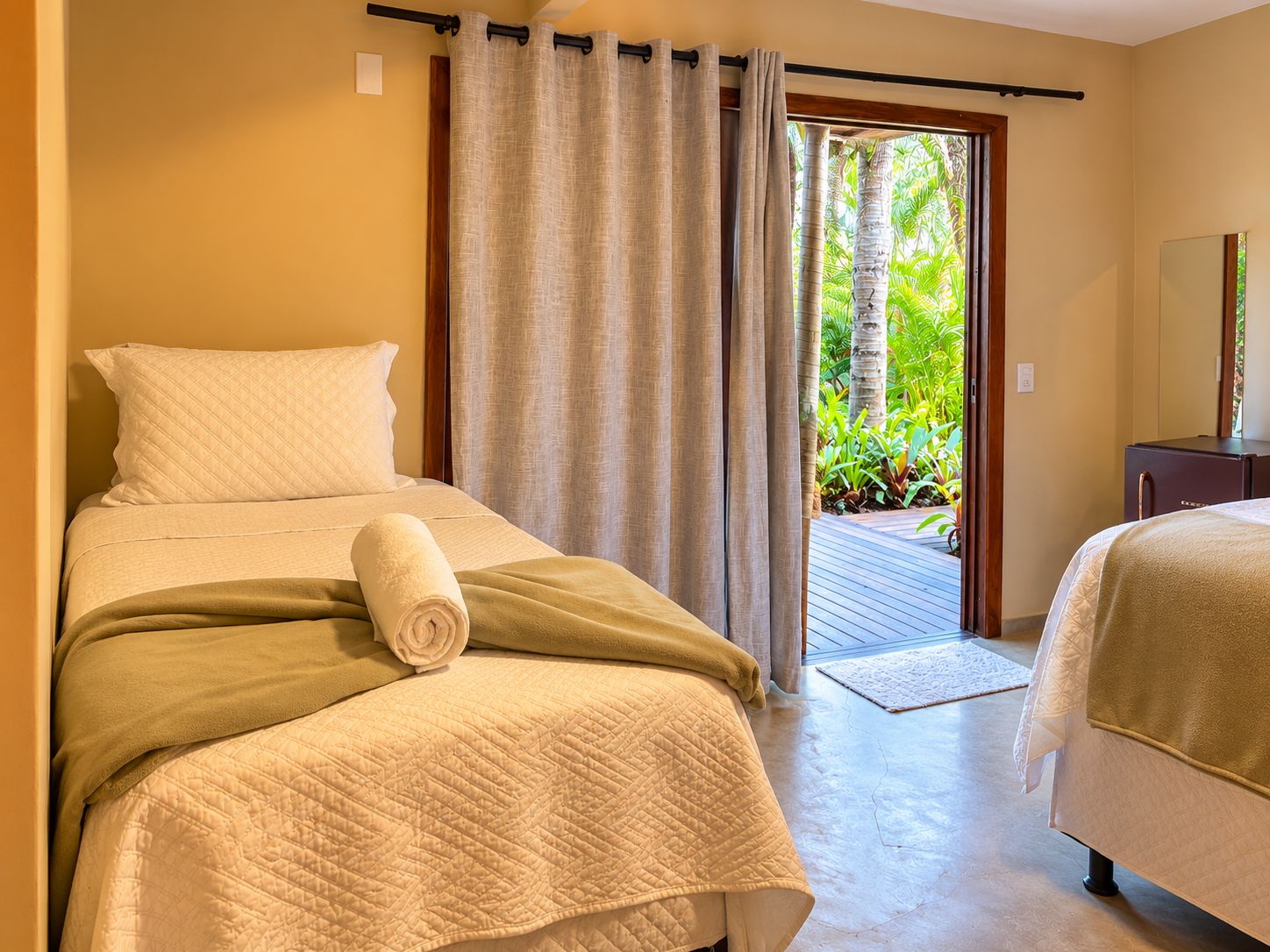 A hotel room with a bed, a rolled towel, a wooden floor, and a view of greenery through the door.