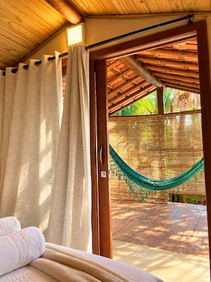 A room with a bed, a hammock, and a view of a wooden deck and greenery.