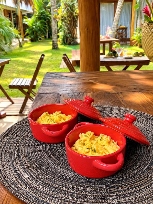 Two red bowls of scrambled eggs on a woven placemat, with a wooden table and chairs, and a tropical garden in the background. - Pousada Jaboticaba Caraíva (Porto Seguro)