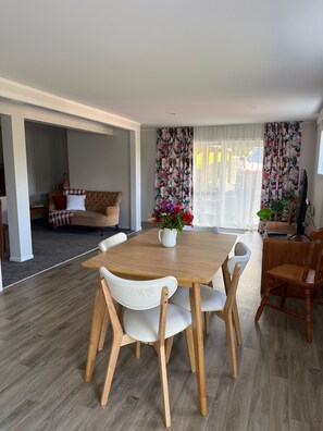 Un comedor con mesa de madera y sillas blancas, una cortina floral y vistas al exterior.