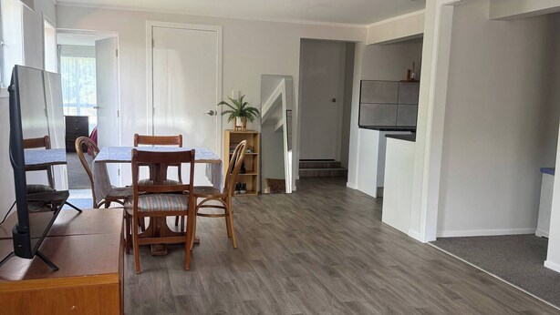 A spacious living room with a wooden floor, a dining area with chairs, a TV on a wooden stand, and a kitchen area with cabinets. - Cozy 2-bedroom House Rental in Whangārei, New Zealand (Whangarei)