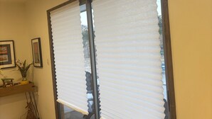 Habitación con puerta corrediza de vidrio, persianas, suelo de madera, una silla y estante con objetos decorativos.