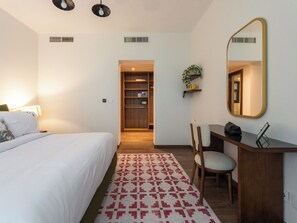 Habitación de hotel con cama, escritorio, silla, espejo, estante con planta y alfombra estampada.