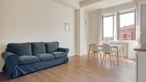 Ein Wohnzimmer mit einer Couch, einem Esstisch mit Stühlen, einem Fenster mit Vorhängen und einer Heizung.