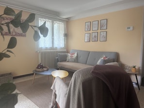 A living room with a sofa, a small table, a radiator, and a window with curtains. - La Soleillade (Manosque)