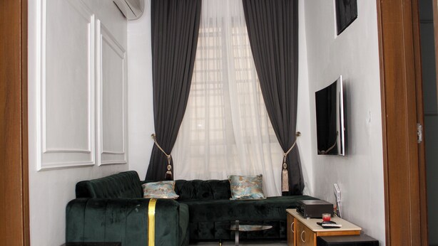 A living room with a dark green sofa, a small side table, a flat-screen TV, and a window with sheer curtains. - Spacious 4-bedroom house in beautiful Lekki with AC and gym (Lekki)