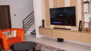 A living room with a large flat-screen TV mounted on a wall, a sofa, a coffee table, and a staircase in the background. - Spacious 4-bedroom house in beautiful Lekki with AC and gym (Lekki)